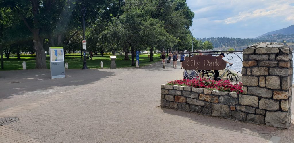 A plaza in the foreground leads to a waterfront walkway in the background with treed parkland to the right.  On the left side of the plaza a stone monument carries a sign that reads 'City Park'