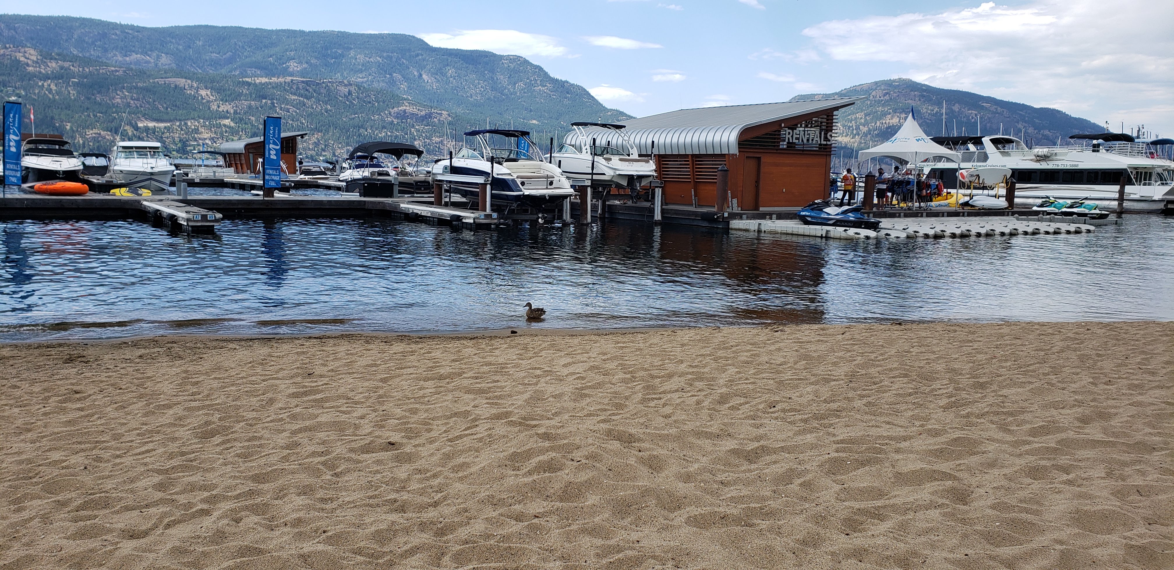 In the foreground a sandy beach. In the middle of the shot boats and docks. In the background a mountain.