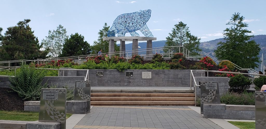 At the end of a wide path, steps and ramps lead to a platform with a piece of public art on it.  A platform atop four pillars carries a blue wire sculpture of a bear, filled with many multi-coloured objects.